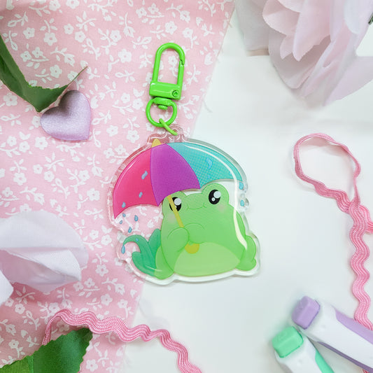 Cute Frog with an umbrella in the rain on an acryllic keychain, features a green clasp and double sided design with epoxy on the front