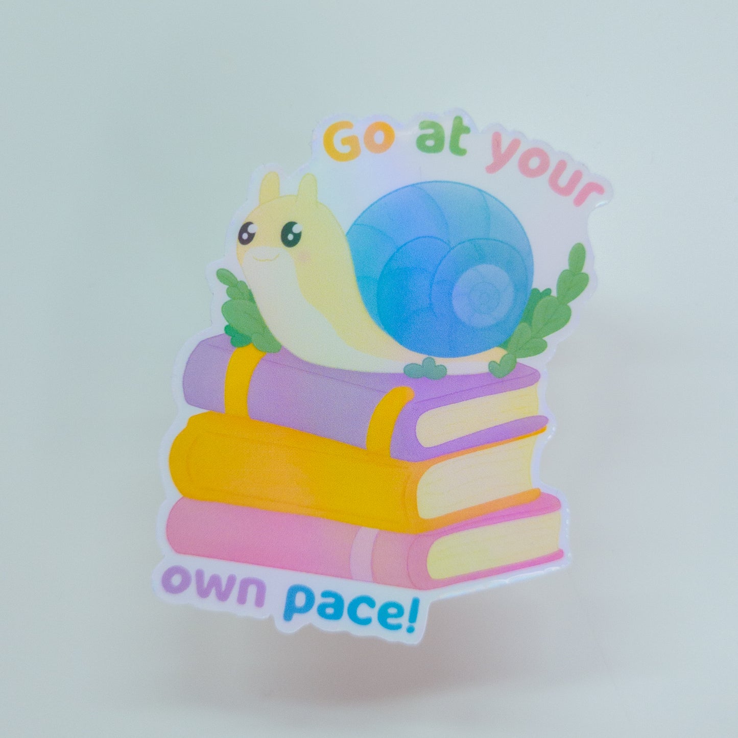 cute bookish sticker, featuring a cute snail on a stack of books and the text 'go at your own pace'
