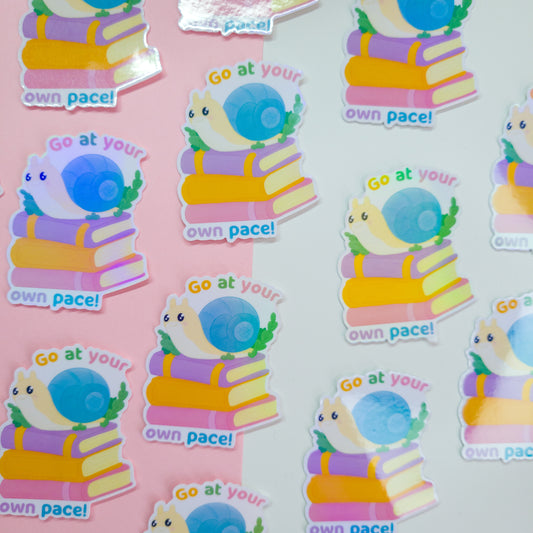 cute bookish sticker, featuring a cute snail on a stack of books and the text 'go at your own pace'