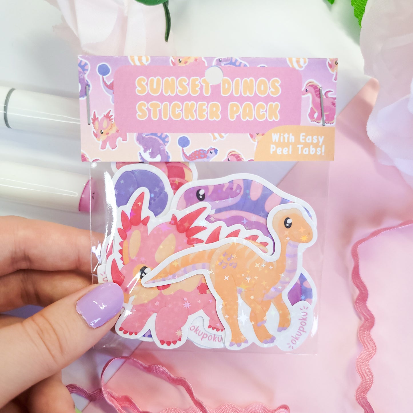 Sticker pack with dinosaur designs held by a hand, against a soft pink background.