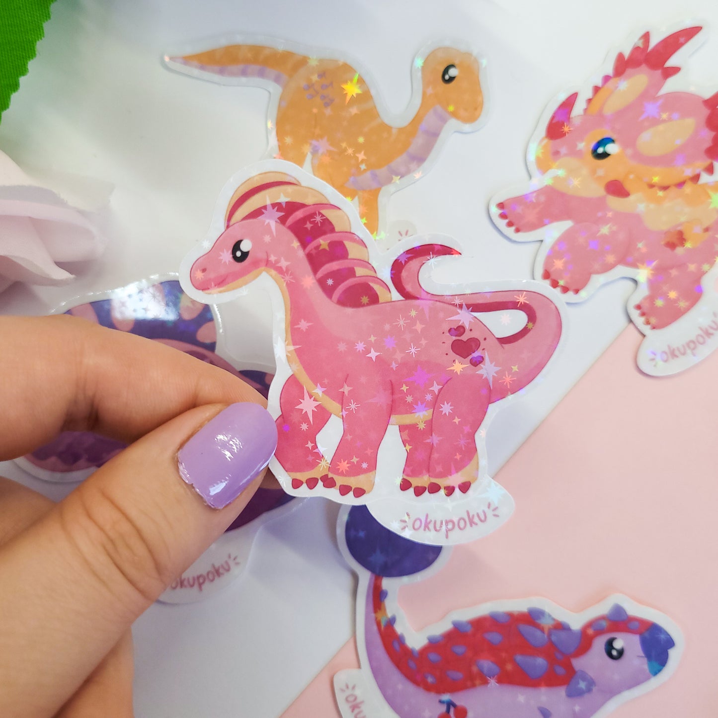 Hand holding a pink amargasaurus dinosaur sticker with other colorful stickers in the background