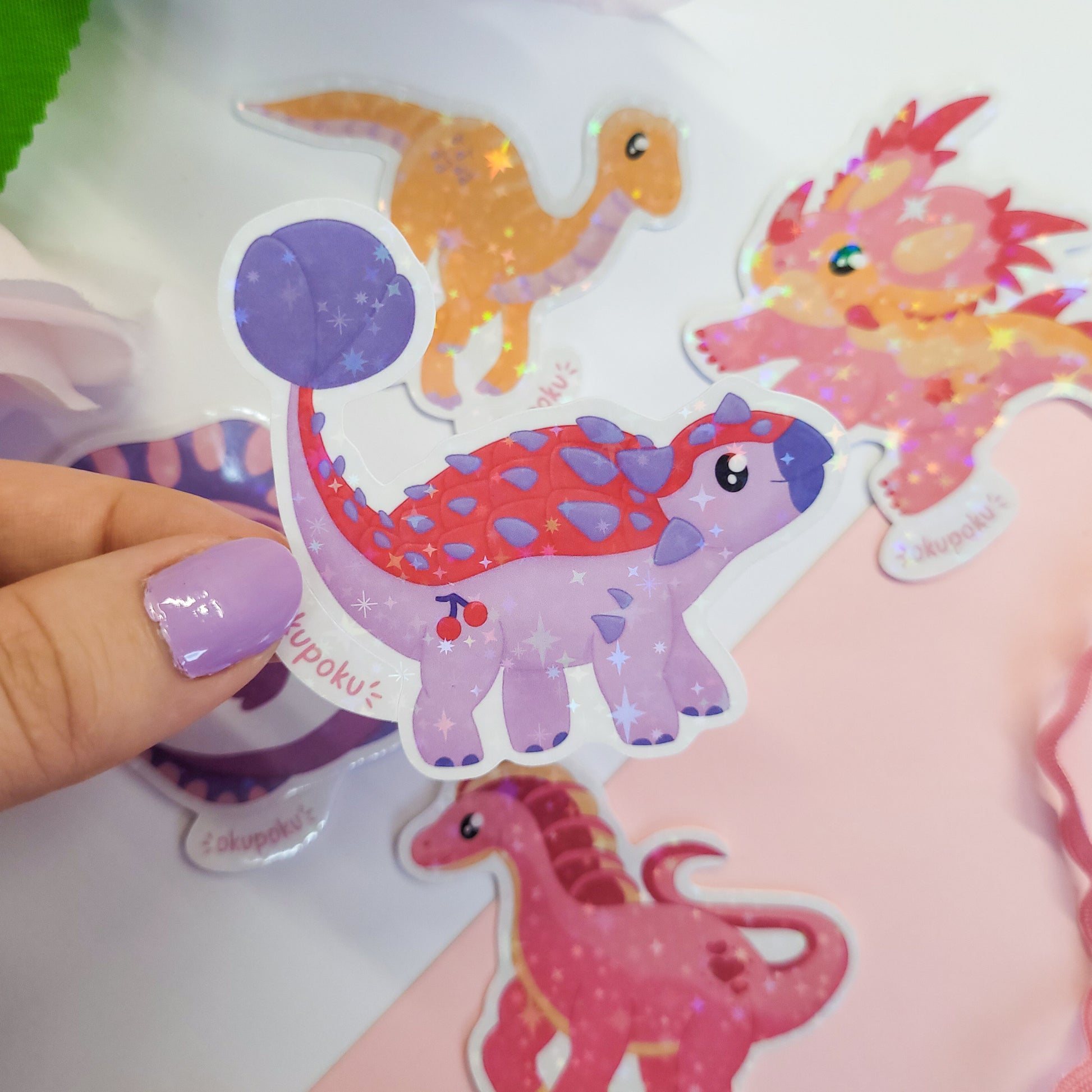Colorful Ankylosaurus dinosaur stickers held by a hand with purple nail polish on a light pink background.